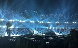 Bright blue spotlights beam across a massive, crowded stadium at night.