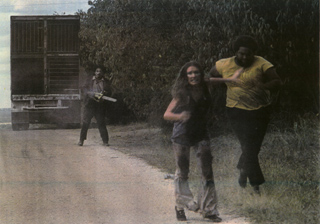 A woman runs down a dirt road while a man with a chainsaw and another man chase her through bushes.