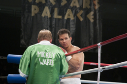 A shirtless boxer looks at his trainer wearing a green robe that says MICKEY WARD in a boxing ring.