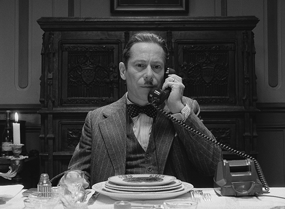 A man in a suit and bowtie speaks into a telephone handset while sitting at a set dining table.