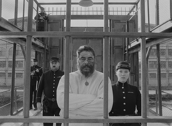 A person in white stands in the foreground with three uniformed guards behind a metal fence.