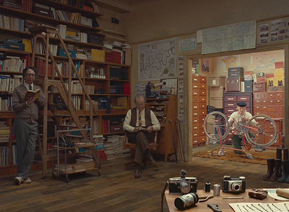 A man reads by a bookshelf, another sits in a chair, and a third works on a bicycle in an adjacent room.