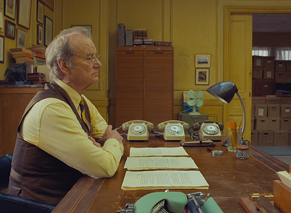 An older man in a yellow shirt and brown vest sits at a wooden desk with papers and rotary phones.