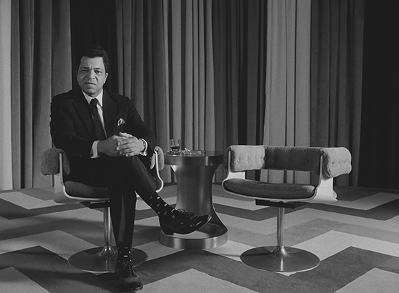 A man in a dark suit sits cross-legged in a modern chair next to a small table and an empty chair.