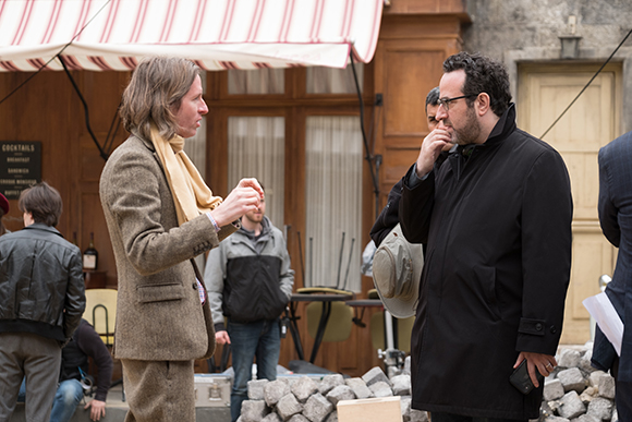 Two men talk outdoors; one wears a tan suit and scarf, the other a black coat.