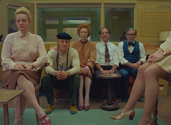 Six people in vintage clothing sit in a room with a map on the wall in the background.