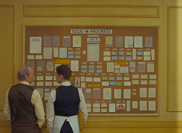 Two men in vests stand before a large brown bulletin board covered in various papers and labels.