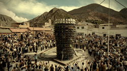 A large crowd of people surrounds a tall, dark, cylindrical structure in a town square with mountains in the background.