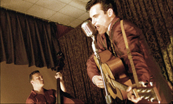 A man in a suit sings into a silver microphone while playing an acoustic guitar on a stage.