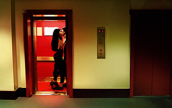 Two people in dark clothing kiss inside a brightly lit red elevator.