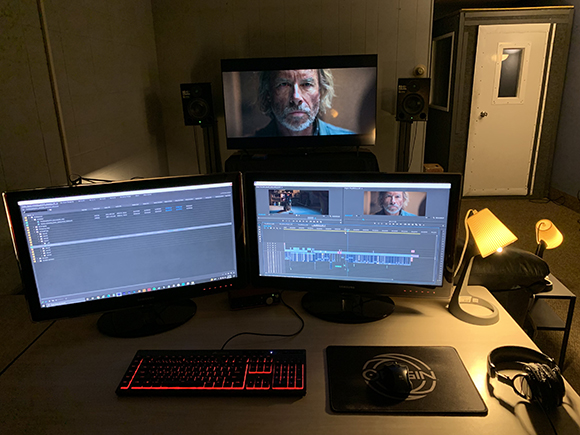Two computer monitors and a TV displaying video editing software and a man's face on a desk with a keyboard and mouse.
