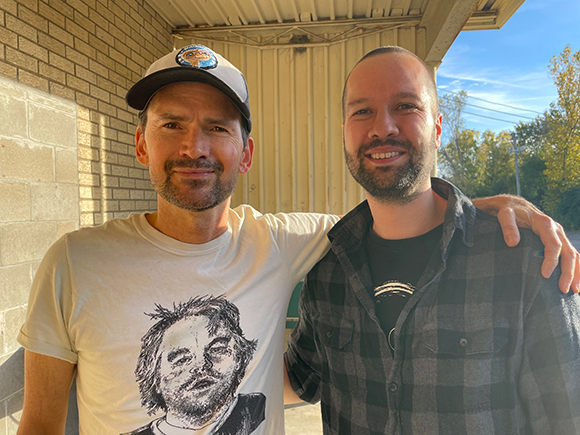 Two men standing side by side, one in a white graphic t-shirt and cap, the other in a plaid shirt.