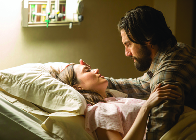 A man with dark hair and a beard leans over a woman lying in a hospital bed, holding her shoulder.
