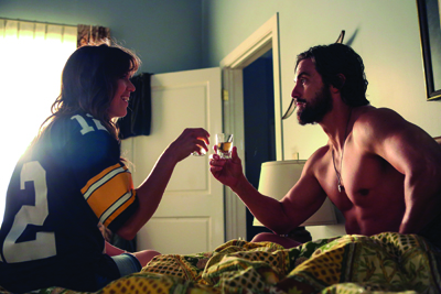 A man and a woman sitting on a bed, smiling and clinking small glasses together.