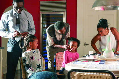 A man, a woman, and three children interact around a dining table in a home.