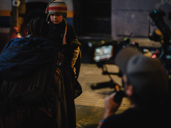 A child in a striped knit hat is held by an adult while a camera crew films in a dimly lit setting.