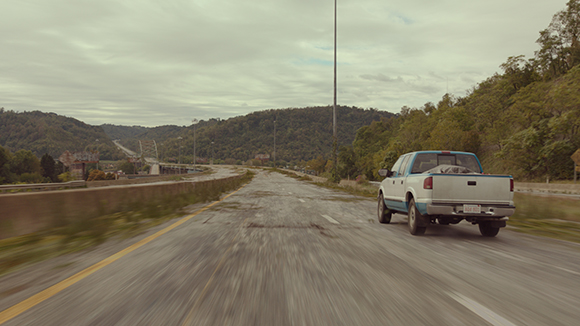 A blue and white pickup truck drives down a multi-lane highway through rolling green hills under a cloudy sky.