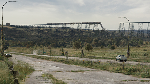 A long, rusted metal trestle bridge stretches across a valley above a cracked, overgrown road.