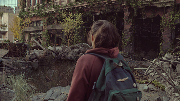 A person wearing a maroon hoodie and green backpack looks toward a crumbling, vine-covered brick building.