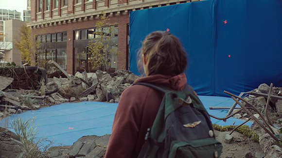 A person with a backpack stands before a large blue tarp and debris near a brick building.
