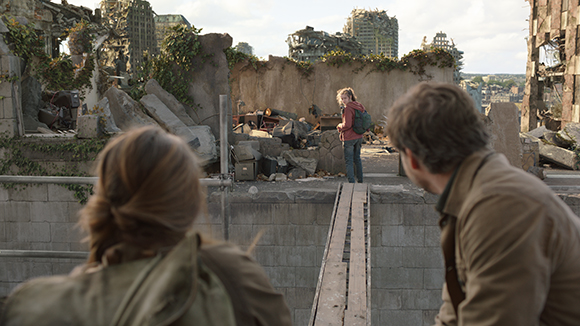 Two people in the foreground look toward a young girl standing in a field of rubble and destroyed buildings.