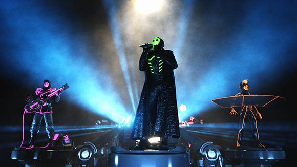 Three performers in glowing, futuristic costumes stand on a dark stage under bright blue spotlights.