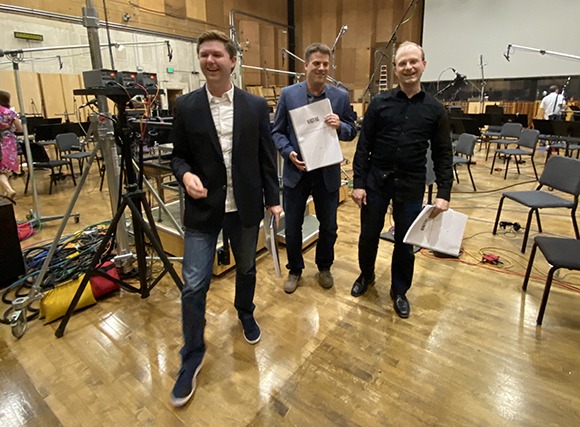 Three men standing together in a large studio with wooden floors and recording equipment.