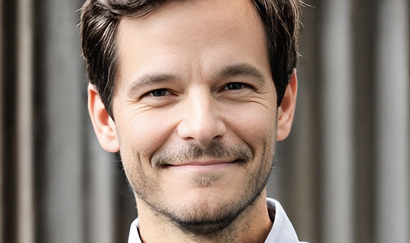 A smiling man with short dark hair and light facial stubble looking toward the camera.