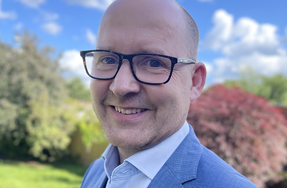 A smiling bald man wearing dark-rimmed glasses and a blue suit jacket over a light blue shirt.