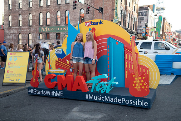 Two young women smiling in front of a large, colorful CMA Fest and US Bank promotional display.