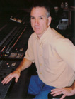 A man in a tan shirt and blue pants sits at a large audio mixing desk.