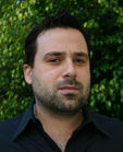 A man with dark hair and facial stubble wearing a black collared shirt against a green leafy background.