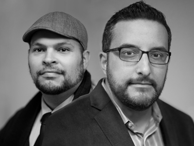 Black and white portrait of two men, one wearing a flat cap and the other wearing glasses and a suit jacket.