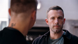 A man with short dark hair and stubble looks forward with a serious expression toward a man seen from behind.