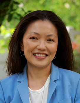 A smiling woman with dark hair wearing a blue blazer and white shirt against a blurred green background.