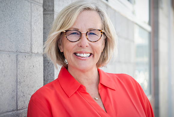 A smiling blonde woman with tortoiseshell glasses wearing a bright red collared shirt.