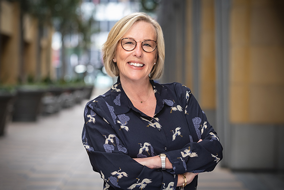 A smiling blonde woman wearing glasses and a navy patterned shirt stands with arms crossed outdoors.