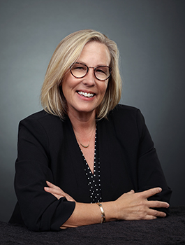 Smiling blonde woman with glasses and a black blazer leaning on a dark surface.