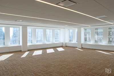 An empty office room with tan carpeting, large windows, and long linear ceiling lights.