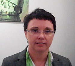 A person with short dark hair and glasses wearing a grey blazer and a green patterned shirt.