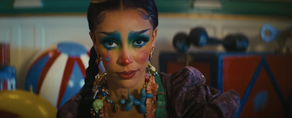 A person with colorful blue and green eye makeup, red nose paint, and long dark braids.