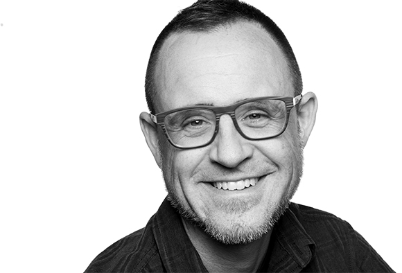 Black and white close-up portrait of a smiling man wearing glasses against a white background.