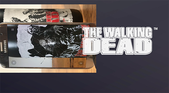 A Samsung smartphone on a wooden surface displaying The Walking Dead artwork with a cracked screen effect.