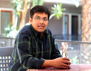 A man wearing glasses and a dark blue and yellow plaid shirt sits at a table outdoors.