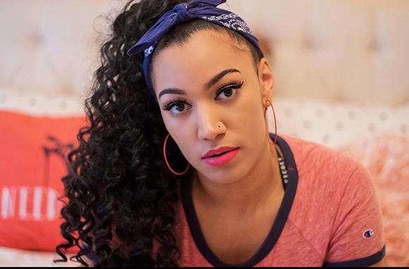 A woman with long curly hair, a blue patterned headband, and pink hoop earrings looks toward the camera.