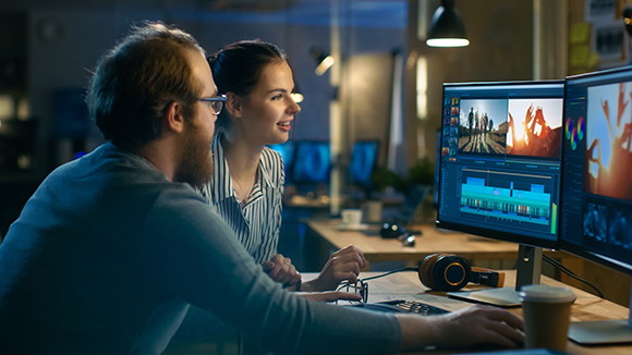 A man and woman look at dual computer monitors displaying video editing software in a dimly lit office.
