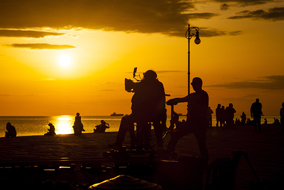 Silhouettes of a film crew and equipment against a bright orange sunset over the ocean.