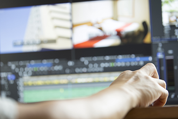 A close-up of a person's hand using a computer mouse to edit video on a multi-screen monitor.