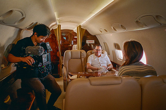 A camera operator films a man and a woman seated at a table inside a private jet.