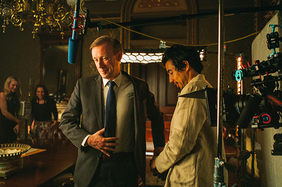 A man in a dark suit and tie stands near a man in a tan jacket on a film set with cameras and lighting.